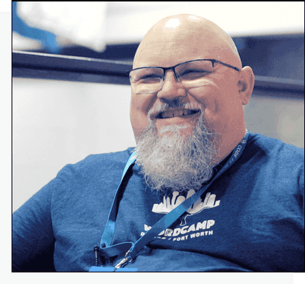 Topher DeRosia wears a WordCamp Dallas / Fort Worth tshirt and smiles, he wears glasses and a grey medium length beard.
