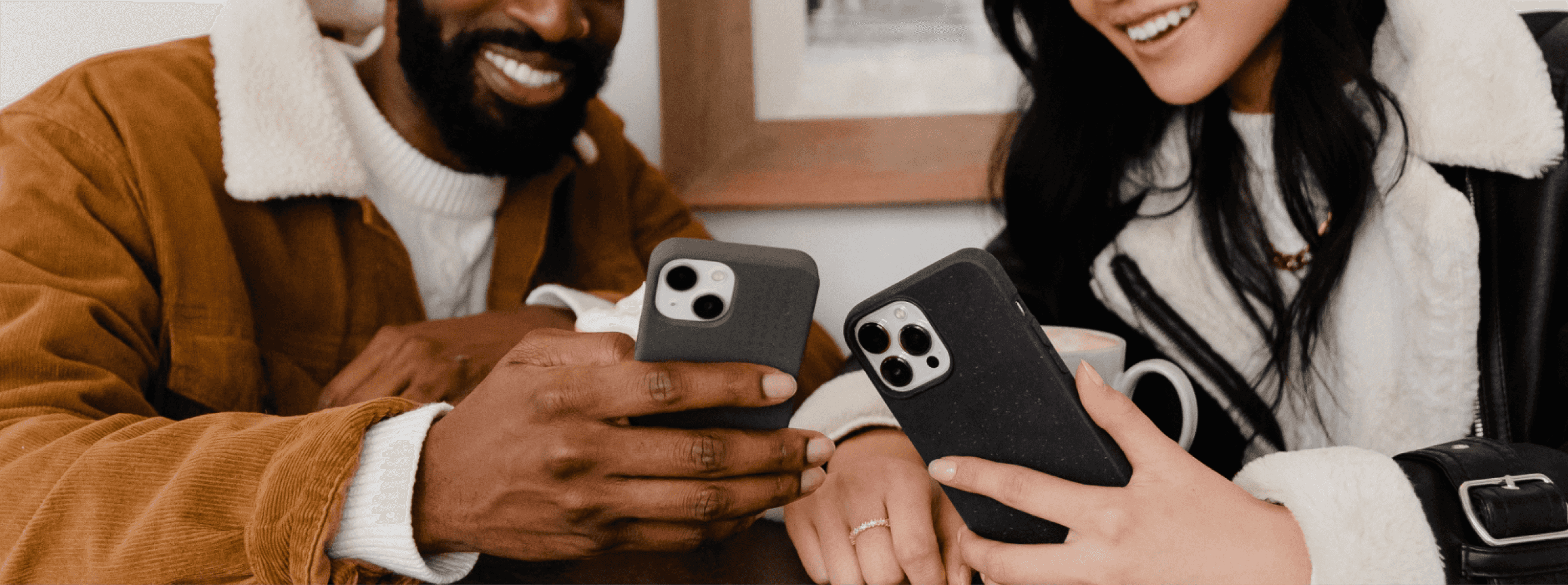 A photo of two people, each holding a phone with a case on it. The background of the photo is a blurred building interior with a framed photo on the wall.