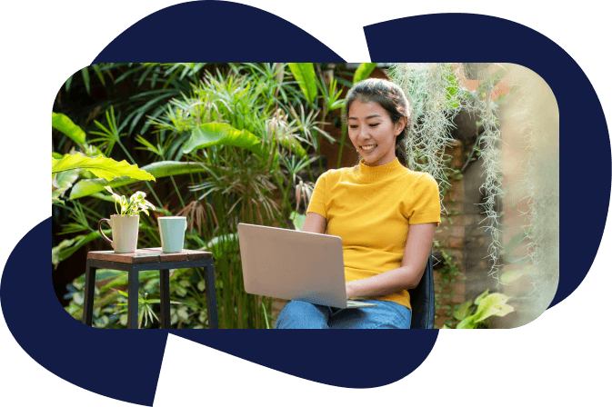 Woman in a turtleneck short sleeve shirt, smiles in a lush garden with 2 coffee cups on a side table, a laptop on her lap